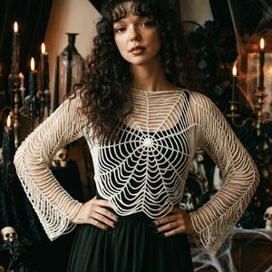 May include: A cream-coloured, long-sleeved crochet top featuring a prominent spiderweb pattern that radiates from the centre of the chest. The garment has a round neckline and flared sleeves, worn over a solid black sleeveless base layer.