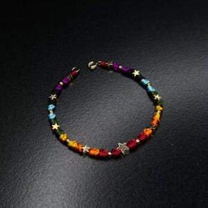 Könnte beinhalten: Ein Armband mit einer mehrfarbigen geflochtenen Kordel in den Farben Rot, Orange, Gelb, Grün, Blau und Violett. Die Kordel ist mit kleinen, goldfarbenen Sternanhängern und winzigen goldfarbenen Perlen verziert und hat einen goldenen Verschluss.