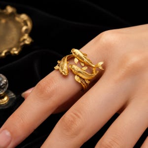 Könnte beinhalten: Ein goldfarbener Ring mit zwei skulptierten Koi-Fischen, die sich um den Finger winden. Die Fische weisen detaillierte Schuppen und Flossen auf, wobei ein Fisch über dem anderen positioniert ist und ein offenes Design bildet, das den Finger umschließt.