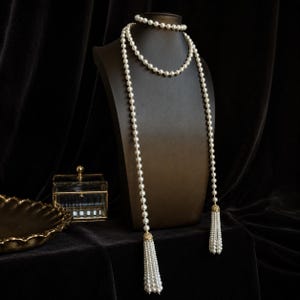 Peut inclure: Un long collier composé d'un rang continu de perles blanches. Le collier est conçu avec deux extrémités pendantes, chacune terminée par un capuchon en métal doré et un pompon de perles à plusieurs brins, créant un aspect superposé.