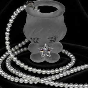 Könnte beinhalten: Eine mattierte Glasfigur in Form einer stilisierten Figur mit großen Augen, die in der Mitte einen klaren, facettierten sternförmigen Kristall aufweist. Die Figur ist an einer langen Schnur aus weißen, kugelförmigen Perlen befestigt.