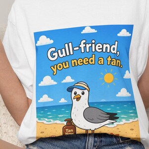 May include: A white t-shirt featuring a graphic of a seagull wearing a visor on a beach with the text Gull-friend, you need a tan. The illustration includes a blue sky with clouds, a yellow sun, the ocean, and a bottle labeled Tan Now.