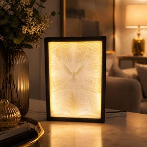 May include: A rectangular light box featuring a warm, glowing relief carving of an open book with a dove soaring above it, surrounded by intricate sunburst rays. The illuminated panel is encased in a simple, thin black frame.