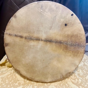 May include: A circular frame drum with a natural, light tan animal hide head. The drum head features a prominent, dark horizontal band across the center and two small, dark circular markings near the upper right edge of the drum surface.