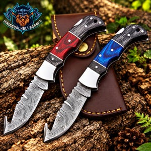 May include: Two folding knives with gut hooks and Damascus steel blades. One knife features a red wood handle, the other a blue wood handle. Both have black wood accents, silver bolsters, and pins. A brown leather sheath is positioned behind them.