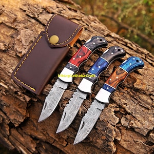 May include: Three folding pocket knives with Damascus steel blades and multi-coloured wooden handles, accompanied by a brown leather sheath with yellow stitching. The text VikingValhallaCraft is visible between the knives and the sheath.