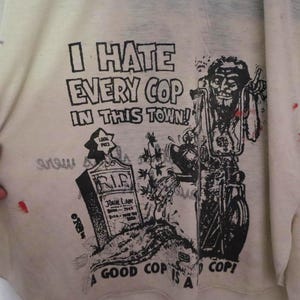 May include: Off-white t-shirt featuring a graphic print with the text "I HATE EVERY COP IN THIS TOWN!". The design includes a tombstone and a biker on a motorcycle, with the phrase "A GOOD COP IS A DEAD COP!" below.