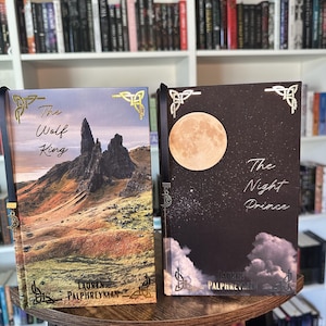 May include: Two hardcover books titled The Wolf King and The Night Prince by Lauren Palphreyman. The Wolf King features a rocky landscape, while The Night Prince shows a full moon in a dark sky. Both books have gold corner accents and bookmarks.
