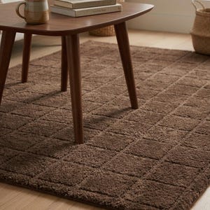May include: A rectangular area rug featuring a deep brown color and a textured, grid-like pattern of raised squares. The rug is placed on a light-colored wooden floor, partially positioned underneath a dark brown wooden coffee table with four legs.