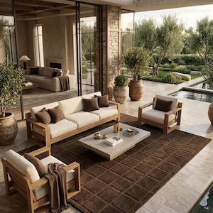 May include: A patio furniture set featuring a light wood sofa and two matching armchairs with cream-colored cushions and dark brown throw pillows. A low rectangular coffee table sits on a large brown grid-patterned rug near a swimming pool.