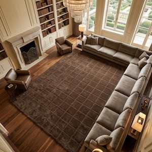 May include: A large, dark brown area rug with a grid pattern covering a wooden floor in a living room. The rug is positioned in the center of a room featuring a large sectional sofa, two leather armchairs, a fireplace, and a chandelier.