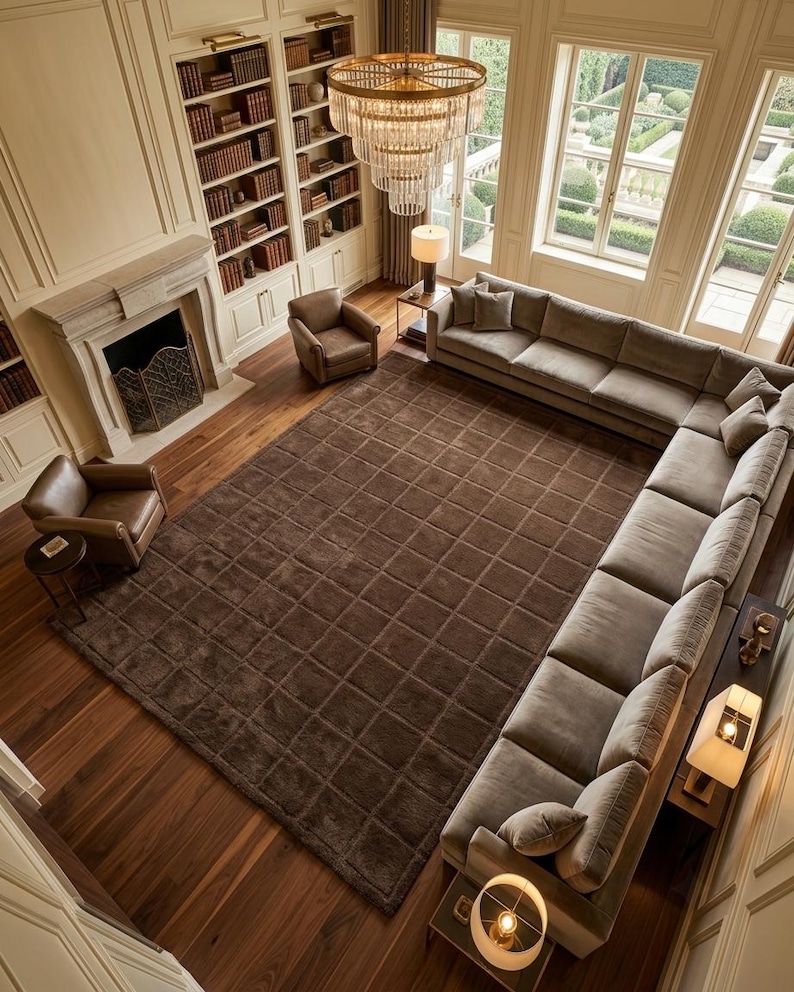 May include: A large, dark brown area rug with a grid pattern covering a wooden floor in a living room. The rug is positioned in the center of a room featuring a large sectional sofa, two leather armchairs, a fireplace, and a chandelier.