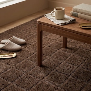 May include: A dark brown square-patterned rug with a pair of beige slippers, a wooden table, a cream-colored ceramic mug, a folded linen napkin, and two stacked books. The rug features a textured, grid-like design across its entire surface.