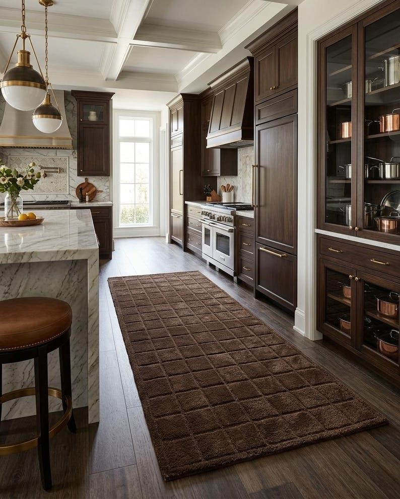 May include: A rectangular runner rug featuring a deep brown color and a raised grid pattern. The rug is positioned on a wooden floor in a kitchen setting, extending lengthwise between a marble-topped island and dark wood cabinetry.
