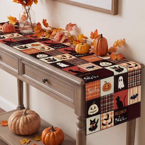 May include: A rectangular table runner featuring a patchwork design of Halloween-themed squares including black cats, ghosts, bats, pumpkins, and the text "Spooky" and "Halloween" in orange, black, white, and plaid patterns, draped over a wooden console table.