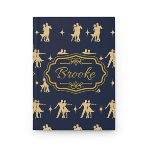 May include: A dark blue hardcover journal featuring a repeating pattern of gold-colored silhouettes of couples dancing. In the center, a decorative gold-colored frame encloses the name Brooke written in a cursive script, surrounded by small stars.