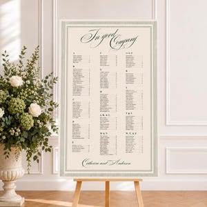 Peut inclure: Un plan de table de mariage rectangulaire sur un chevalet en bois avec un fond crème et du texte vert foncé. L'en-tête indique In good Company, suivi d'une liste alphabétique de noms et de numéros de table, finissant par Catherine and Anderson.
