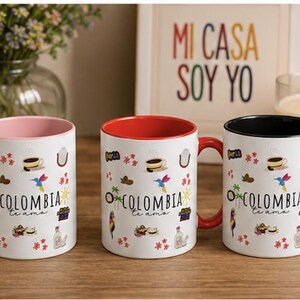 May include: Three white ceramic mugs featuring the text COLOMBIA te ama and colorful illustrations of a hummingbird, coffee, and tropical elements. Each mug has a distinct colored handle and interior rim in light pink, red, and black respectively.