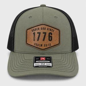 May include: An olive green trucker hat with a black mesh back and a brown hexagonal leather patch on the front. The patch features the black text "UNDER GOD SINCE 1776" and "PSALM 33:12". A black and white Richardson 112 sticker is on the brim.