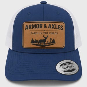 Könnte beinhalten: Eine blau-weiße Trucker-Cap mit einem beigefarbenen Patch in Lederoptik auf der Vorderseite. Der Patch zeigt den Text ARMOR & AXLES, FAITH IN THE FIELDS und eine Grafik eines Hirsches auf einem Feld. Ein runder Aufkleber auf dem Schirm zeigt THE AUTHENTIC YUPOONG SNAPBACK.