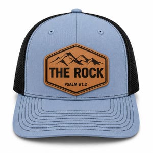 May include: A light blue baseball cap with a black mesh back and a brown hexagonal leather patch on the front. The patch features a black mountain graphic, the text THE ROCK, and the scripture reference PSALM 61:2 in black lettering.