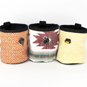 May include: Three colorful climbing chalk bags with geometric patterns. The bags are made of fabric and have a drawstring closure. Each bag has a metal clip on the side.