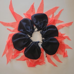 May include: Five black drawstring pouches arranged in a circular pattern, each featuring a black cord with a plastic toggle closure. The pouches are layered over a bright neon orange fabric base with multiple pointed, petal-like fabric strips.