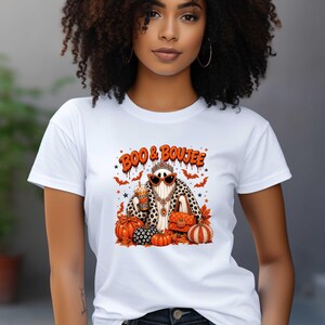 May include: A white short-sleeved t-shirt featuring a graphic of a ghost wearing a leopard print coat, sunglasses, a tiara, and a necklace. The ghost holds a drink and a handbag, surrounded by orange pumpkins, bats, and the text BOO & BOUJEE.