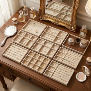 May include: A collection of beige velvet jewelry organizer trays arranged on a wooden surface, featuring various compartments for rings, necklaces, and earrings, accompanied by perfume bottles, a hand mirror, and a gold-framed wall mirror.