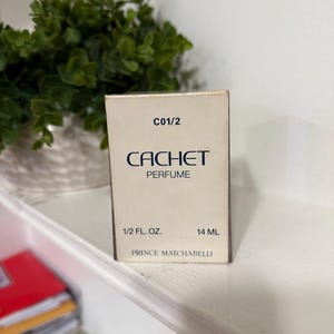 May include: A rectangular off-white perfume box with the text C01/2, CACHET PERFUME, 1/2 FL. OZ., 14 ML, and PRINCE MATCHABELLI printed in dark blue and black lettering on the front face of the packaging.