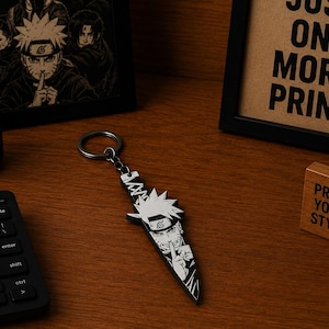 May include: A black and white kunai-shaped keychain featuring a detailed graphic illustration of the character Naruto Uzumaki from the anime series Naruto. The keychain includes a metal ring and a chain attachment, showcasing a sharp, pointed design.
