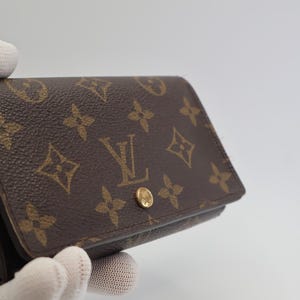 May include: A brown leather wallet featuring the iconic Louis Vuitton monogram pattern in a tan color. The rectangular wallet has a fold-over flap secured by a small, circular gold-toned metal snap button closure centered on the front exterior.