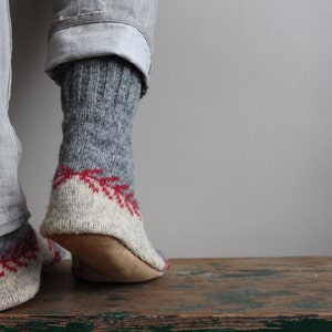 May include: A pair of gray wool socks with red and white knit patterns. The socks are worn and the feet are resting on a wooden surface.