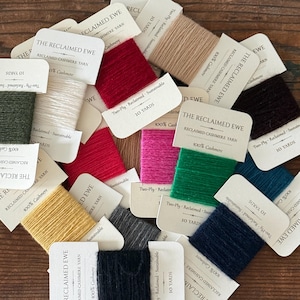 May include: A collection of small cards wrapped with 10 yards of two-ply reclaimed cashmere yarn in various colors including red, green, yellow, white, black, and blue. Each card is labeled with The Reclaimed Ewe and 100% Cashmere text.