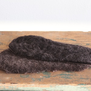 May include: Two dark brown, fluffy insoles are displayed on a weathered wooden surface. The insoles are oval-shaped, designed to fit inside shoes, and appear to be made of a soft, textured material, possibly wool or a similar fabric.