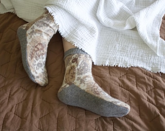 May include: A pair of ankle-length socks featuring a cream and brown paisley pattern with solid grey heels, toes, and cuffs. The socks are worn under a white textured blanket on a brown quilted surface, showcasing a soft, knit-like fabric texture.
