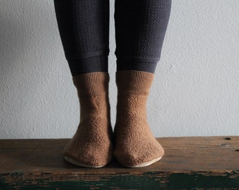 May include: A pair of brown fuzzy socks with a ribbed texture. The socks are worn on a person's feet and are resting on a wooden surface.