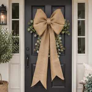 May include: A large, tan burlap bow with long, hanging tails centered on a wreath of green eucalyptus leaves. The bow is constructed from a coarse, woven fabric and features two wide loops and two long, pointed tails that drape downward.