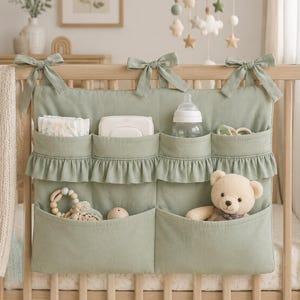 May include: A sage green fabric crib organizer featuring three top pockets with ruffled trim and two larger bottom pockets. The organizer is attached to a wooden crib rail with three fabric ties. It holds diapers, wipes, a bottle, and a teddy bear.