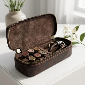 May include: A dark brown rectangular zippered travel case with a soft suede-like interior lining. Inside, the case contains five small circular metal pins featuring the letters C, O, R, D, and B, alongside a braided dark brown leather bracelet.