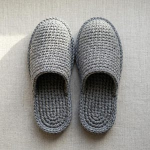 Puede incluir: Un par de zapatillas de color gris claro confeccionadas con un patrón de ganchillo denso y texturizado. Las zapatillas presentan un diseño de punta redondeada y una abertura superior, mostrando una técnica de punto uniforme.
