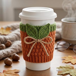 May include: A white reusable coffee cup featuring a textured orange crochet sleeve with a ribbed green top edge. The sleeve is decorated with two green crocheted leaves and a central tan twine bow tied in a knot, creating a pumpkin-like appearance.