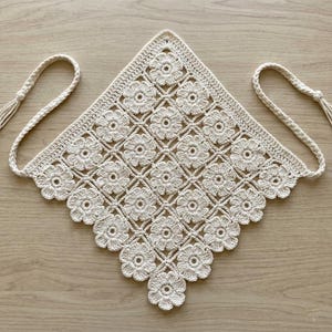 May include: A diamond-shaped off-white crochet bandana featuring a repeating pattern of interconnected floral motifs. The piece is constructed with intricate yarn stitching and includes two long braided ties extending from the top corners.