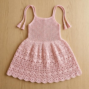 Pode incluir: Um vestido sem mangas rosa claro feito com técnicas de crochê. O corpete apresenta um padrão de grade vazado, enquanto a saia exibe um design de renda denso e intrincado inspirado em flores. Duas alças finas trançadas com borlas pendem para baixo.