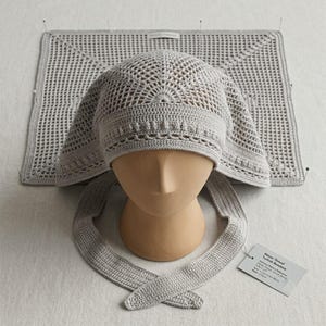 May include: A light gray crocheted head covering with a delicate openwork pattern and long ties, displayed on a mannequin head. A matching rectangular crocheted piece lies flat behind it, and a small white tag with printed text is attached.