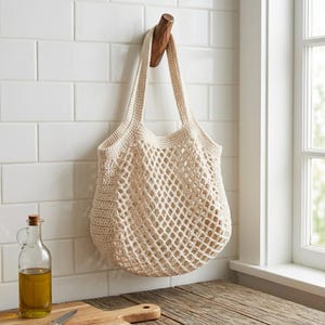 Peut inclure: Un sac de marché en crochet de couleur crème présentant un motif en maille en forme de losange au centre et un point de crochet plein le long du haut, des côtés et des anses. Le sac est suspendu à un crochet en bois sur un mur carrelé blanc.