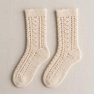 May include: A pair of cream-colored socks crafted with a detailed crochet knit pattern. The socks feature a scalloped edge at the top cuff, a vertical chevron-style openwork design running down the leg, and a solid knit heel and toe section.