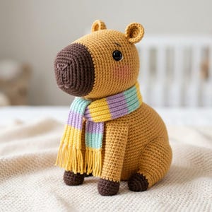 May include: A crocheted capybara stuffed animal with golden-brown yarn, a dark brown snout, and black plastic eyes. It wears a striped scarf featuring pastel yellow, light blue, and lavender colors with fringe at the ends. The feet are dark brown.