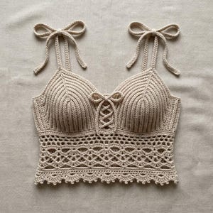 May include: A beige crochet crop top featuring a ribbed bodice with a central lace-up detail, two shoulder straps that tie into bows at the top, and a decorative openwork pattern along the bottom hem with a scalloped edge finish.