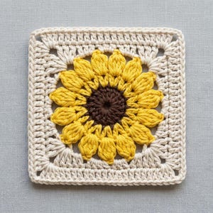 May include: A square crochet motif featuring a central sunflower design with a dark brown center and bright yellow petals. The sunflower is framed by a cream-colored border worked in a classic square pattern with visible stitches and texture.